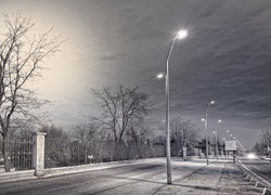 Czarno-białe zdjęcie ulicy nocą z latarniami i ogrodzeniem