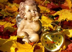 Figurka aniołka obok zegara w kształcie serca w jesiennych liściach