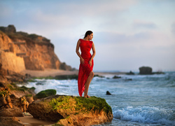 Kobieta w czerwonej sukience pozuje na kamieniu przy brzegu morza