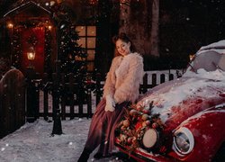 Kobieta w futerku oparta o ośnieżony samochód w świątecznej scenerii