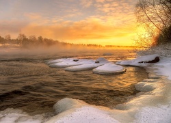 Zachód słońca nad oblodzoną rzeką i zamglonymi zimowymi drzewami