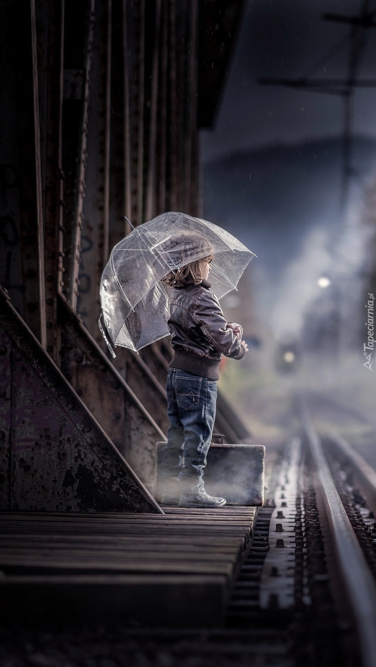 Dziecko z walizką pod parasolem czekające na pociąg