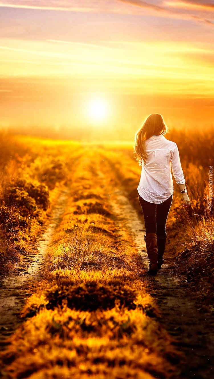 Kobieta idąca polną drogą ku zachodzącemu słońcu