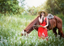 Azjatka w letniej sukience i kapeluszu obok pasącego się konia