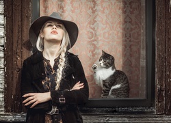 Blondwłosa dziewczyna w kapeluszu i kotek za oknem