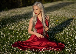 Blondynka w czerwonej sukience siedzi na łące pośród stokrotek