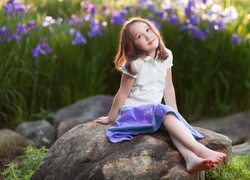 Bosonoga dziewczynka siedzi na kamieniu z irysami w tle