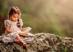 Bosonoga dziewczynka z warkoczami przegląda książkę siedząc na kamieniu