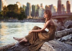 Bosonoga kobieta w sukience siedzi nad brzegiem rzeki