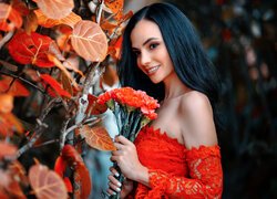 Brunetka w czerwonej sukni z goździkami w rękach obok jesiennych liści na gałęziach
