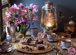 Bukiet kwiatów w wazonie obok lampy oraz filiżanek z kawą i ciasta