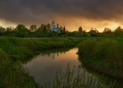 Cerkiew wśród drzew nad rzeką pod ciemnymi chmurami