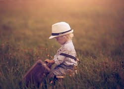 Chłopczyk z walizką w kapeluszu pośród traw