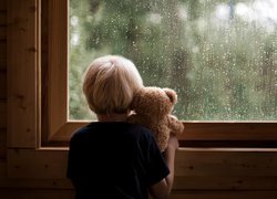Chłopiec z pluszowym misiem patrzący przez okno w kroplach deszczu