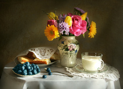 Ciasto, borówki i filiżanka mleka obok wazonu z kwiatami na stoliku