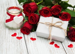 Czerwone róże obok prezentu przewiązanego sznurkiem i palącej się świecy