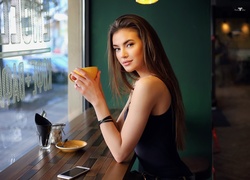 Długowłosa kobieta z filiżanką w dłoniach siedzi w kawiarni przy oknie
