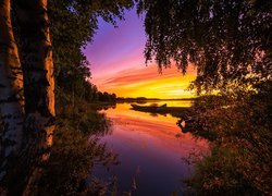 Drzewa i łódka na brzegu jeziora pod kolorowym niebem zachodzącego słońca