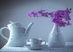 Dzbanek i filiżanka obok wazonu z kwiatami za mokrą szybą