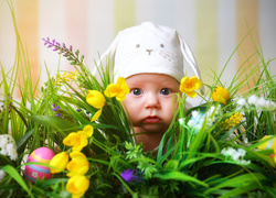 Dziecko w czapeczce spogląda zza wielkanocnej kompozycji kwiatowej z pisankami
