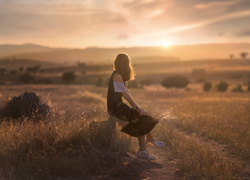 Dziewczyna pośród uschniętych traw wpatrzona w zachodzące słońce