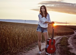 Dziewczyna z gitarą na polnej drodze w promieniach zachodzącego słońca
