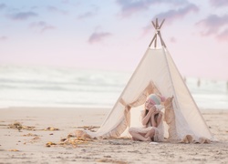 Dziewczynka przed namiotem rozbitym na plaży słucha szumu morza wydobywającego się z muszli