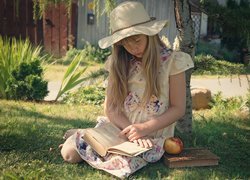 Dziewczynka w kapeluszu czytająca książkę pod drzewem