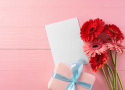 Gerbery i prezent obok pustej kartki na różowych deskach