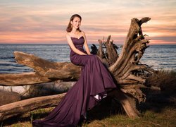 Kobieta w długiej sukni siedząca na suchym konarze drzewa nad morzem