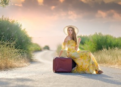 Kobieta w zwiewnej letniej sukience i kapeluszu na drodze z walizką