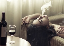 Kobieta wydmuchująca dym z papierosa i butelka wina z kieliszkiem na stole