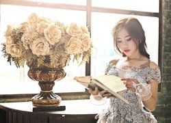 Kobieta z książką obok róż w wazonie przy oknie