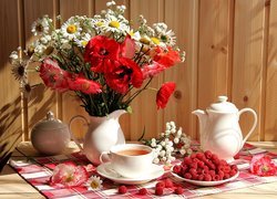 Kwiaty w dzbanku obok herbaty w filiżance i malin na talerzyku