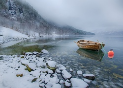 Łódka na kamienistym brzegu jeziora na tle zimowych gór i drzew
