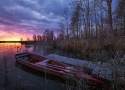 Łódka przy pomoście oraz szuwary i bezlistne drzewa nad jeziorem o zachodzie słońca