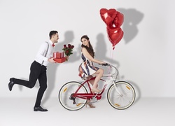 Mężczyzna z prezentem i kwiatami biegnący za kobietą na rowerze