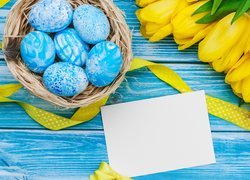 Niebieskie pisanki w koszyczku obok żółtych tulipanów
