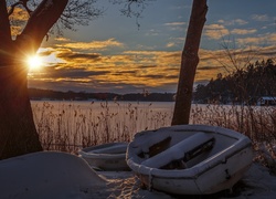 Ośnieżone łódki pod drzewami na brzegu jeziora o zachodzie słońca