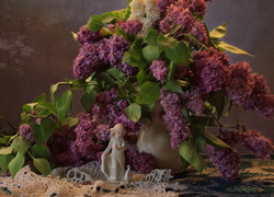 Porcelanowa figurka stoi na serwetce obok wazonu z kwiatami bzu