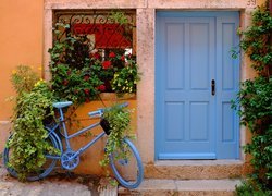 Rower z kwiatami pod oknem obok niebieskich drzwi