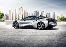 Srebrne BMW i8 rocznik 2014 z drapaczami chmur i drzewami w tle