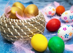 Złote jaja w koszyczku obok kolorowych pisanek i piór