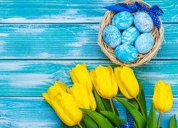 Żółte tulipany obok koszyczka z niebieskimi pisankami na deskach